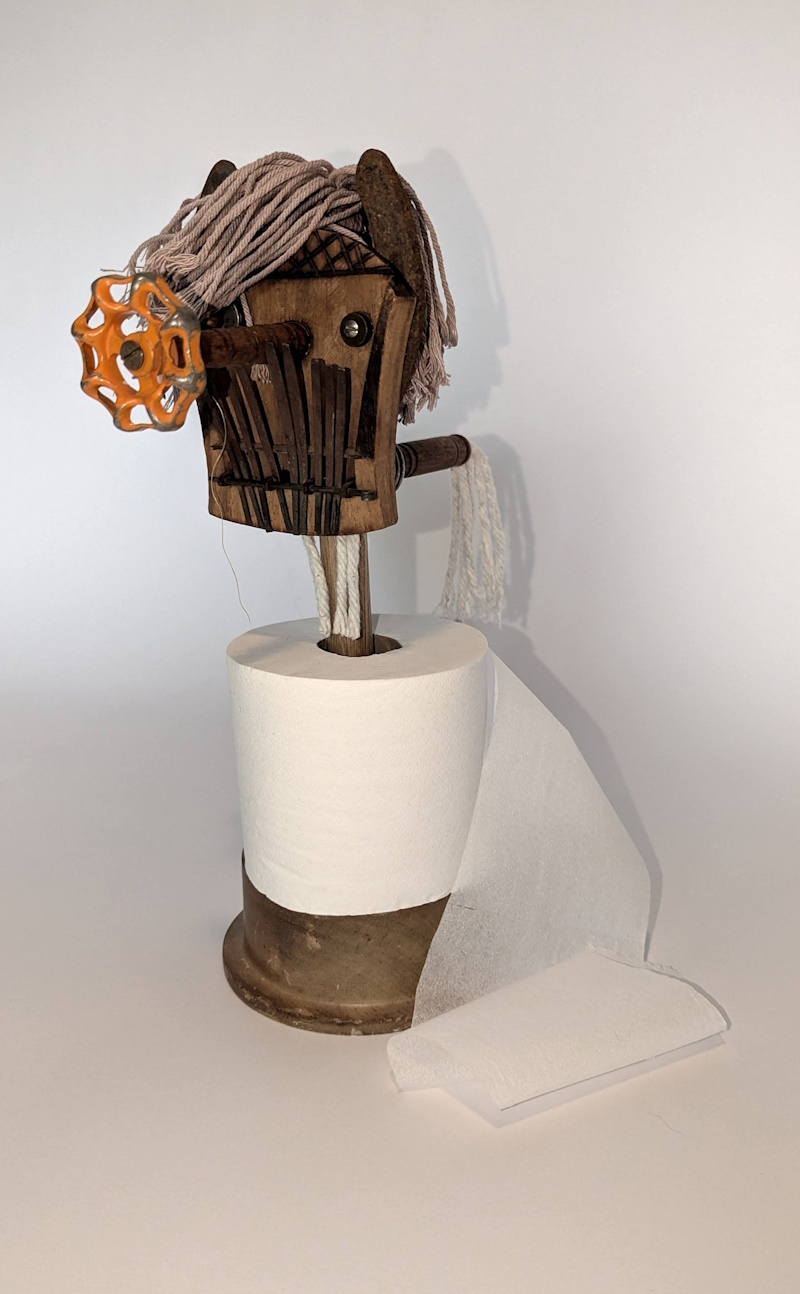 A whimsical assemblage sculpture named Hairy Harry, featuring a quirky figure with a playful expression, creatively holding a toilet paper roll on a stand. The piece blends humor and functionality, showcasing various materials in a fun and unconventional design.