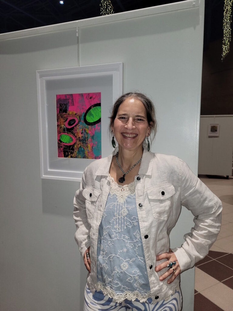 Artist standing beside a framed monoprint with collage at The Liminal Print solo exhibition, featuring 70 works at the Atrium Gallery in 2026.