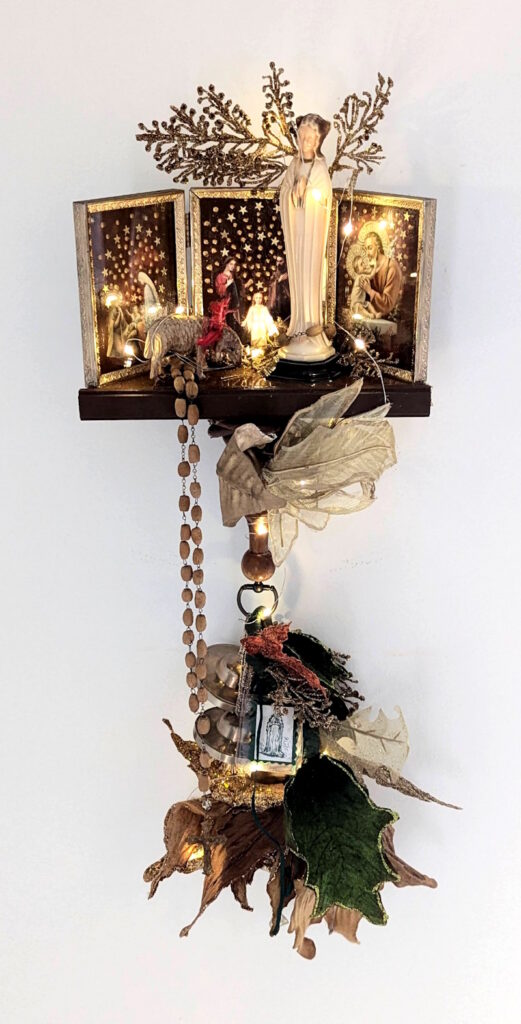 : Illuminated wall-mounted assemblage shrine featuring a central Virgin Mary figure, glowing lights, religious imagery, rosary beads, leaves, metallic branches, bells, and layered found objects against a white background.