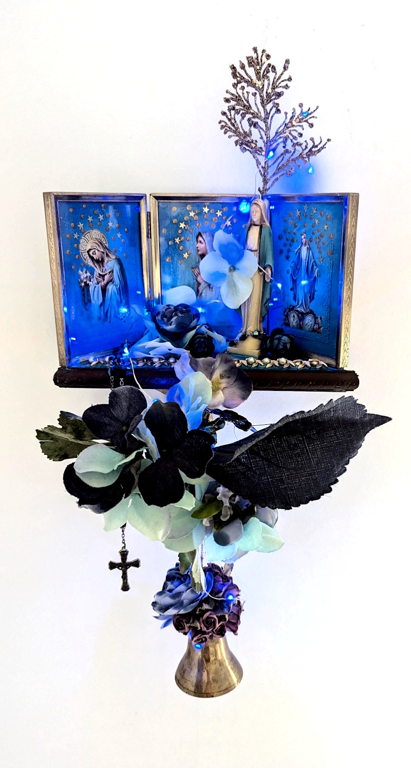 Illuminated wall-mounted assemblage shrine with Marian imagery, blue LED lights, flowers, a hanging cross, coral-like branches, and a brass bell against a white background.
